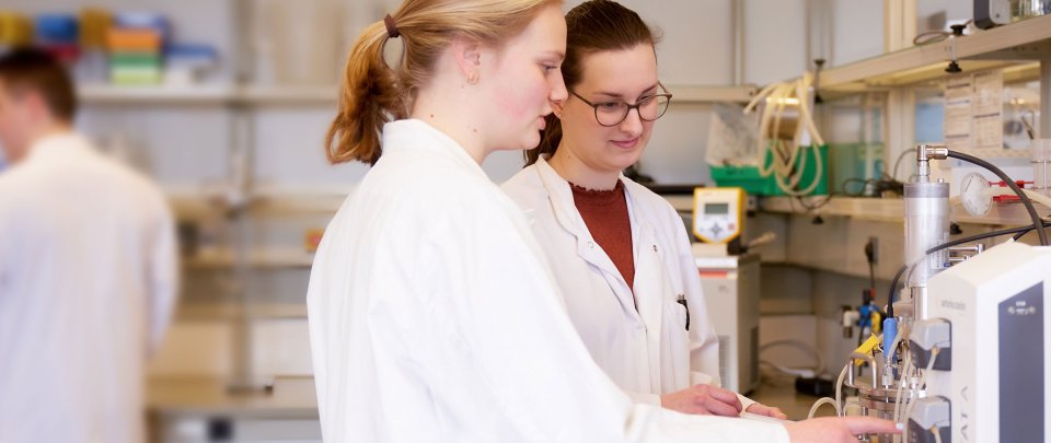 Zwei junge Frauen im Labor. Sie stehen an einer Anlage und füllen etwas in einen Glasbehälter. Sie tragen Laborkittel.