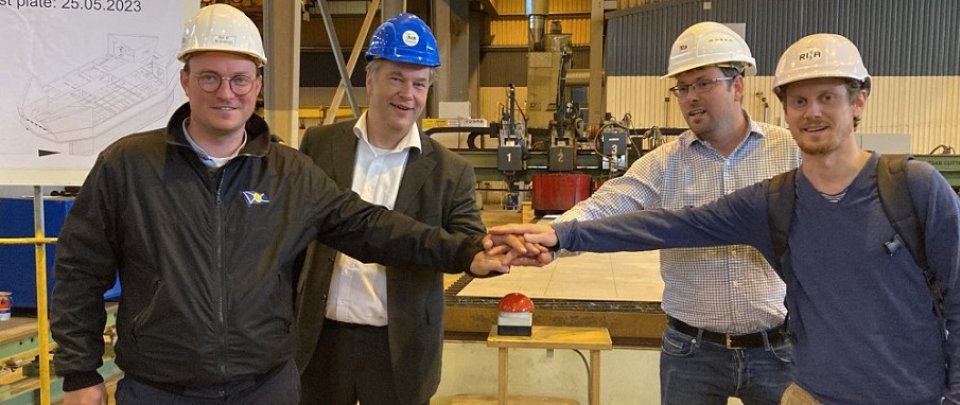 Vier Männer stehen in einer Werfthalle rund um einen großen roten Knopf. Sie haben die Hände übereinander auf den Knopf gelegt und lächeln in die Kamera. Alle vier tragen Bauschutz-Helme.