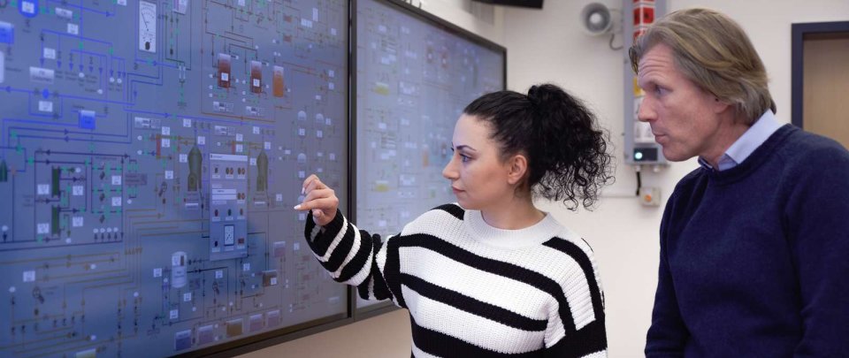 Eine Studentin und ein Dozent stehen vor einer Reihe von großen, an der Wand aufgehängten Monitoren. Auf den Monitoren sieht man eine schematische Darstellung der verschiedenen Gerätschaften in einem Schiffs-Maschinenraum. Die Studentin zeigt auf etwas, der Dozent schaut aufmerksam zu.