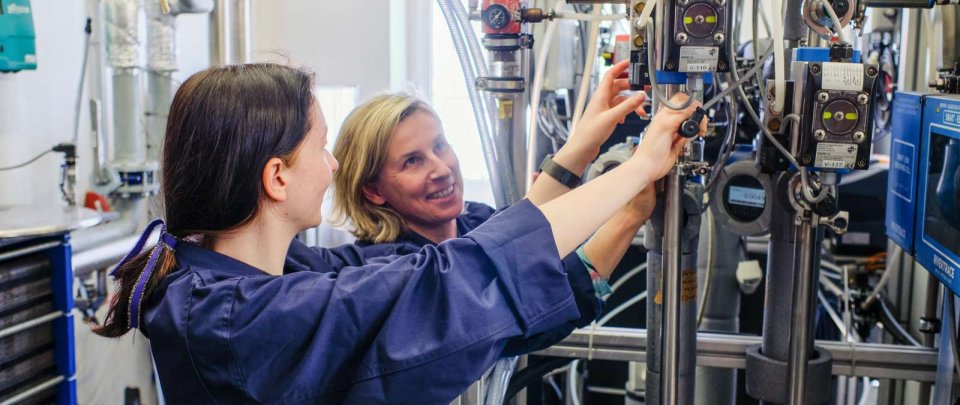 Eine Studentin und eine Dozentin arbeiten lächelnd gemeinsam an einer mehr als kopfhohen, leuchtend blauen Anlage. Beide tragen dunkelblaue Arbeits-Schutzanzüge, über ihren Kopf laufen verschiedene Schläuche und Rohre von der Maschine.