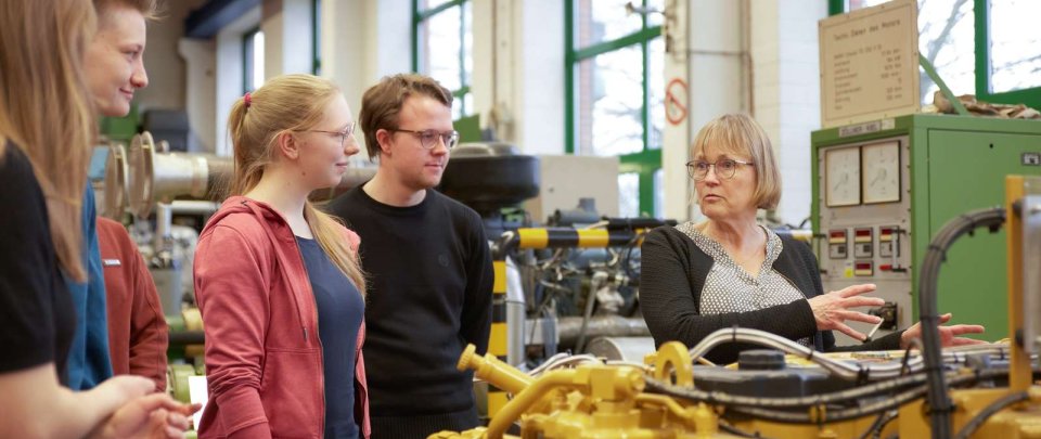 Eine Dozentin, zwei Studentinnen und zwei Studenten stehen um einen leuchtend gelben Motorblock. Die Dozentin, klein mit blonden Haaren und Brille, gestikuliert beim Erklären, die Student*innen blicken sie aufmerksam an.