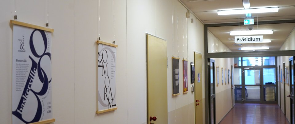 Ein Flur, in dem mehrere Poster mit künstlerisch gestalteter Schrift an der Wand hängen. Mittig im Flur hängt ein Glasschild mit der Aufschrift "Präsidium".