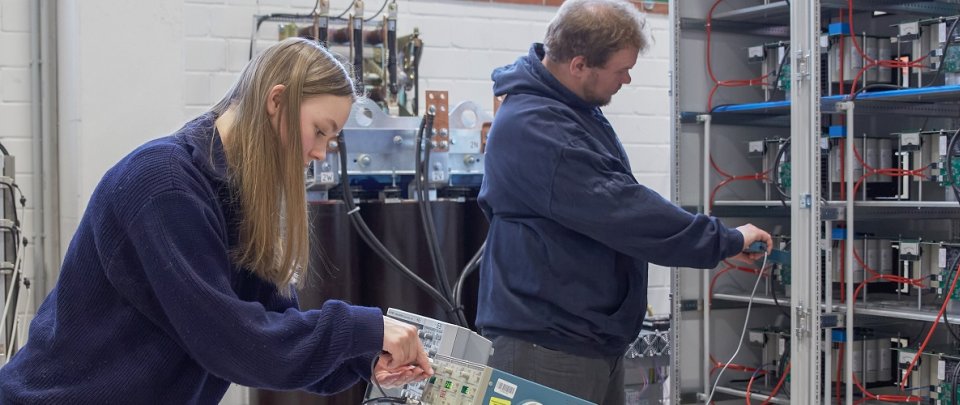 Eine Studentin und ein Student stehen an einer elektrischen Schaltvorrichtung und stecken verschiedene Kabel um.