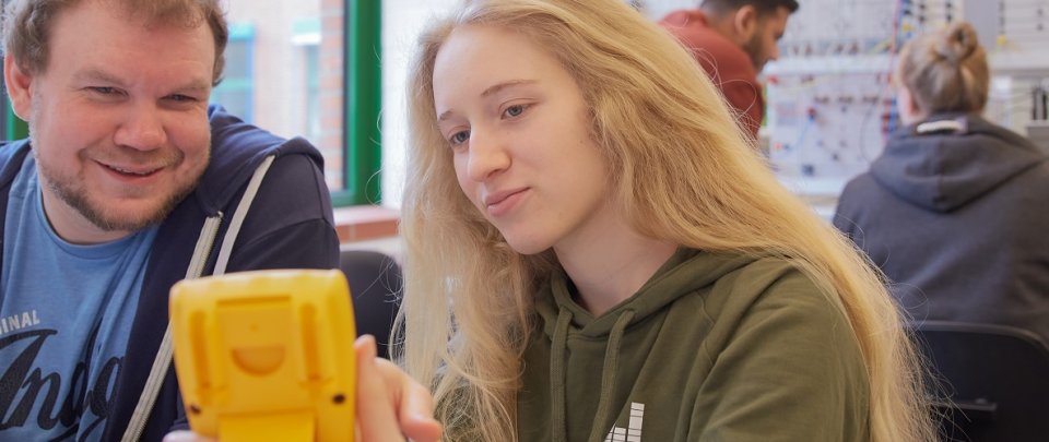 Ein Student und eine Studentin blicken gemeinsam auf ein gelbes Messgerät, das die Studentin in der Hand hält. Im Hintergrund sieht man zwei Studierende an einer elektrischen Schaltvorrichtung hantieren.