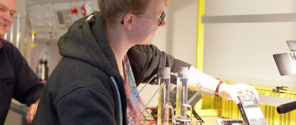 Eine junge Frau hält ein Messgerät unter eine Leuchte, daneben steht eine Elektrolyse-Apparatur. Neben ihr sitzt ein älterer Mann und beobachtet ihre Arbeit aufmerksam.