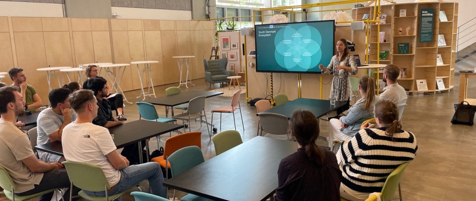 Eine Gruppe von Studierenden sitzt in einem weitläufigen, in hellem Holz getafelten Raum; vor ihnen steht eine Frau vor einem Monitor, auf dem ein Kreisdiagramm zu sehen ist mit der Überschrift "South Denmark Ecosystem".