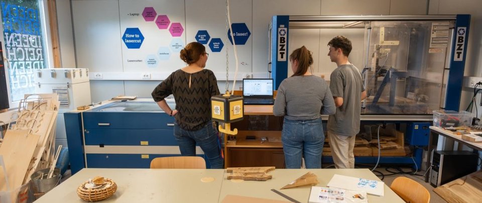 Studierende im ideenreich. Im Bildvordergrund ein Arbeitstisch mit Lineal, Notizbuch, Spanplatten, im Hintergrund ein Lasercutter.