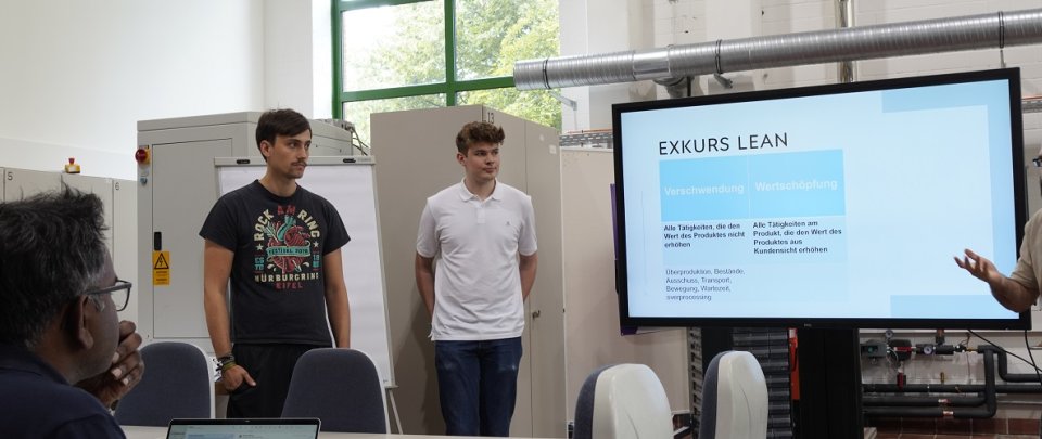 Eine Gruppe von vier Studierenden steht um einen großen Monitor , einer von ihnen spricht gestikulierend mit zwei Dozenten, die vor ihnen an einem Tisch sitzen.