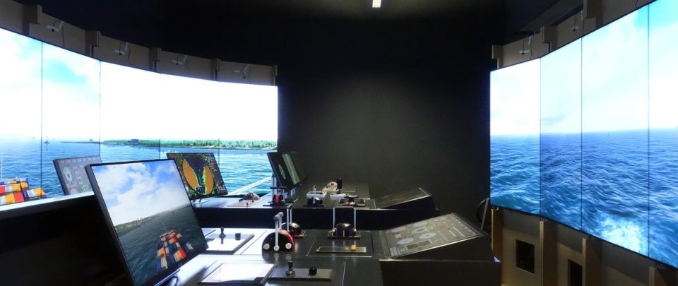 Ein dunkler Raum, in dem vorne und hinten große Monitore stehen, auf denen eine Hafenumgebung zu sehen ist, in der Mitte ein Pult mit Monitoren und Steuervorrichtungen wie auf einer Schiffsbrücke.