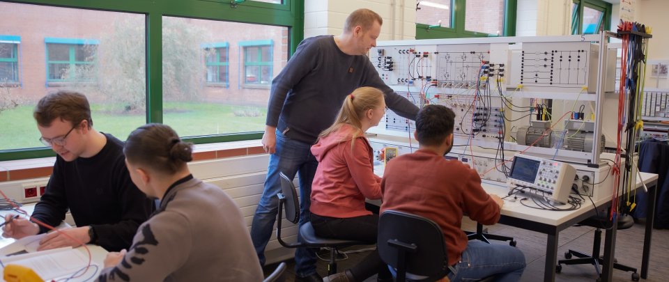 In einem Laborraum sitzen Studierende an Tischen, auf denen aufwändige elektrische Schaltvorrichtungen aufgebaut sind. Ein Dozent steht hinter zwei Studierenden und zeigt auf etwas an der Schaltwand.