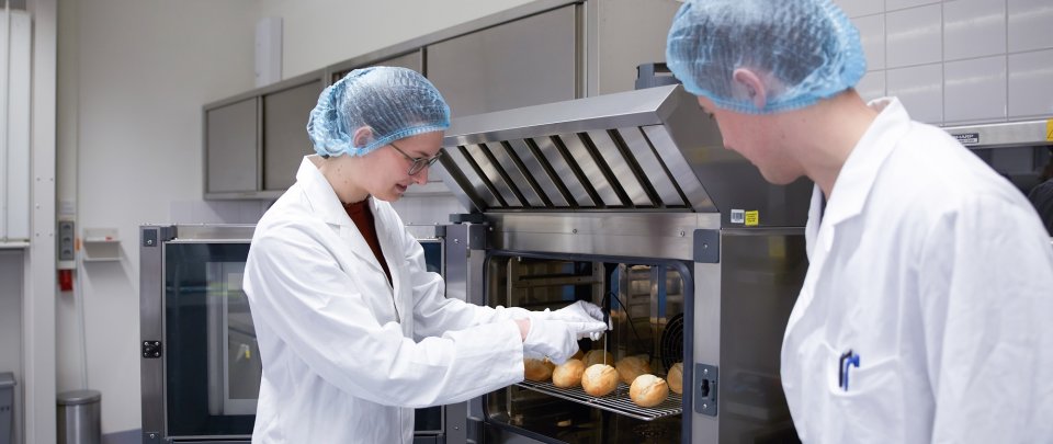 In einem Laborraum stehen zwei Studierende in Laborkitteln an einem Backofen und prüfen mit einem Thermometer die Temperatur in einem Brötchen.