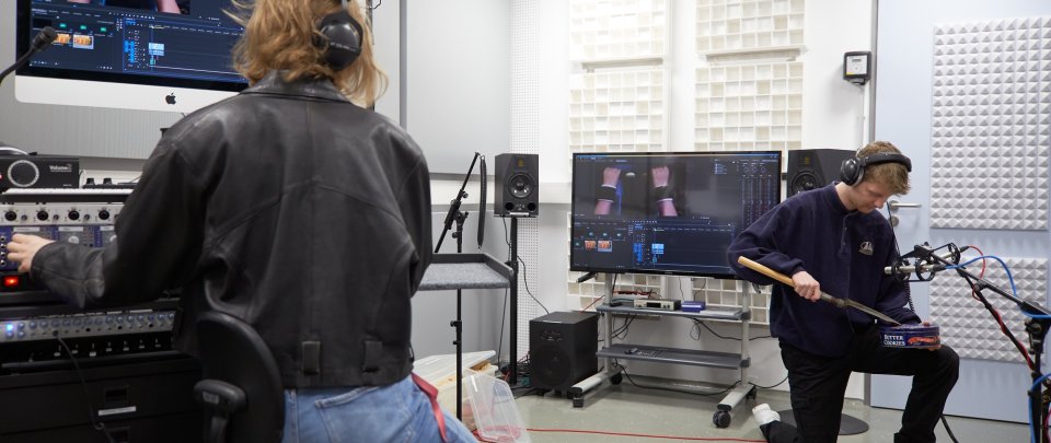 In einem Studioraum sitzt eine junge Frau in schwarzer Lederjacke an einem Mischpult, über dem ein Monitor aufgebaut ist, an dem ein Videobearbeitungsprogramm zu sehen ist. Ein junger Mann kniet vor einem Mikrofon und reibt mit einem Metallspaten über eine Keksdose.