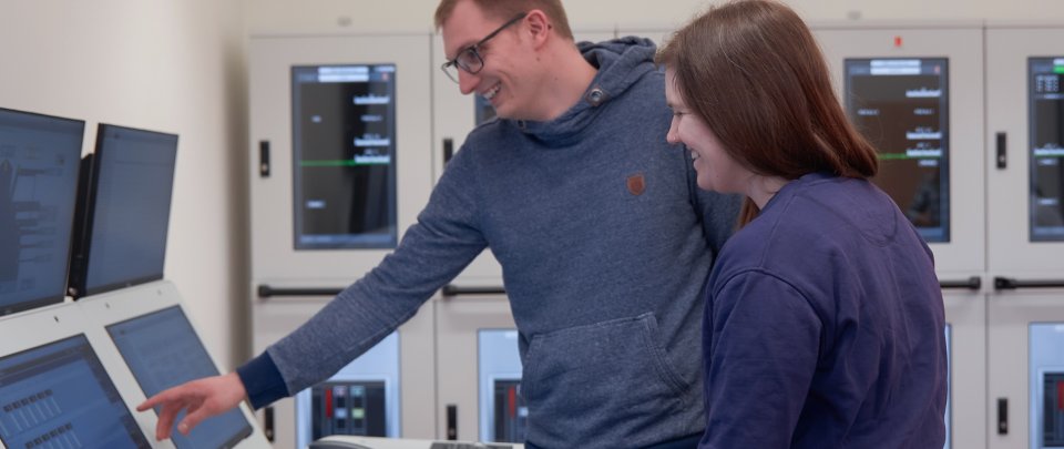 Ein Student und eine Studentin stehen in einem Raum mit vielen Monitoren, auf denen schiffsbetriebstechnische Anzeigen zu sehen sind.