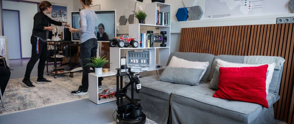 In einem Raum mit Sofa, Teppichen, Regal und Schreibtisch steht im Vordergrund ein Roboterarm auf dem Boden, im Hintergrund stehen zwei Frauen vor einem großen Monitor und deuten darauf.