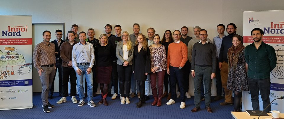 Eine Gruppe von mehr als 20 Menschen steht in einem Raum und lächelt in die Kamera. Rechts und links neben ihnen stehen zwei Roll-Ups mit Text und Bildern unter der Überschrift Inno!Nord.