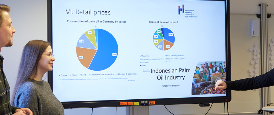 In einem Seminarraum stehen ein Student und zwei Studentinnen vor einem großen Monitor, auf dem verschiedene Tortendiagramme zu sehen sind - neben einem steht in größerer Schrift "Indonesian Palm Oil Industry".