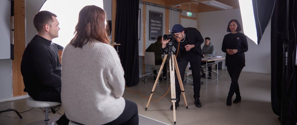 In einem Raum sitzen zwei Studierende auf Hockern und werden angestrahlt, ein weiterer Student macht mit einer Kamera auf einem Stativ Fotos von ihnen, während eine Dozentin in schwarzer Kleidung neben ihm steht.