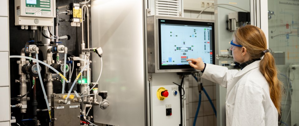 Eine Frau mit langem blondem Zopf im Laborkittel steht vor einer technischen Anlage mit vielen Schläuchen und Anzeigen und tippt auf einen Monitor.