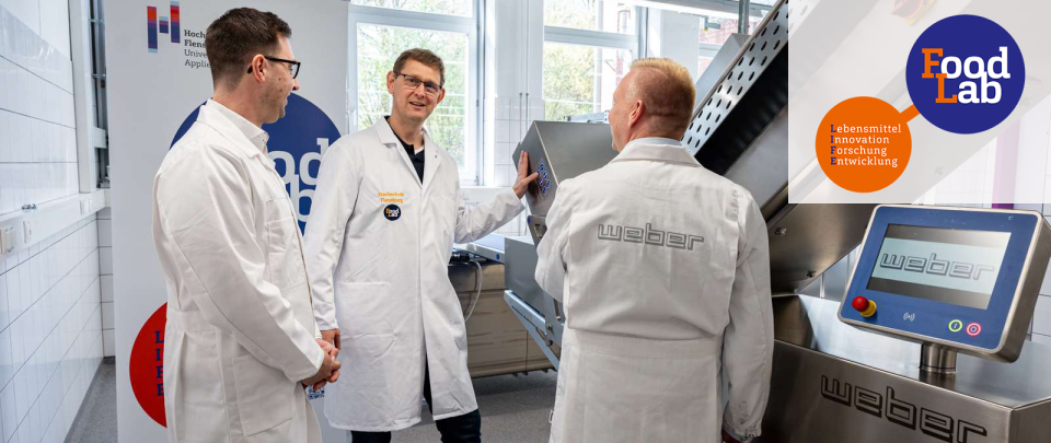 Drei Männer mittleren Alters in Laborkitteln stehen an einem mehr als mannshohen silbernen Laborgerät mit der Aufschrift "Weber".