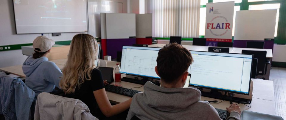 In einem PC-Laborraum sitzen drei Studierende mit dem Rücken zum Fotografen an PC-Arbeitsplätzen. Im Hintergrund eine Leinwand mit einer Präsentation und ein Roll-Up mit dem Schriftzug FLAIR und dem Logo der Hochschule Flensburg.