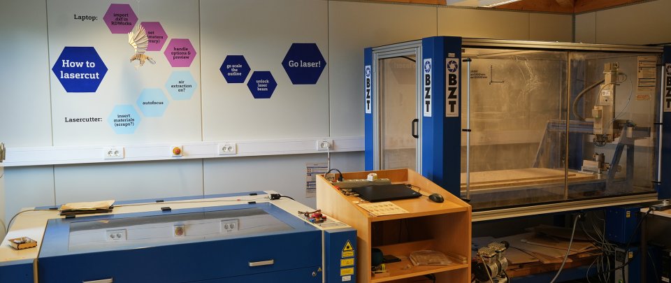 In einem Raum stehen ein Lasercutter und eine CNC-Fräse, an der Wand sind bunte Rauten.