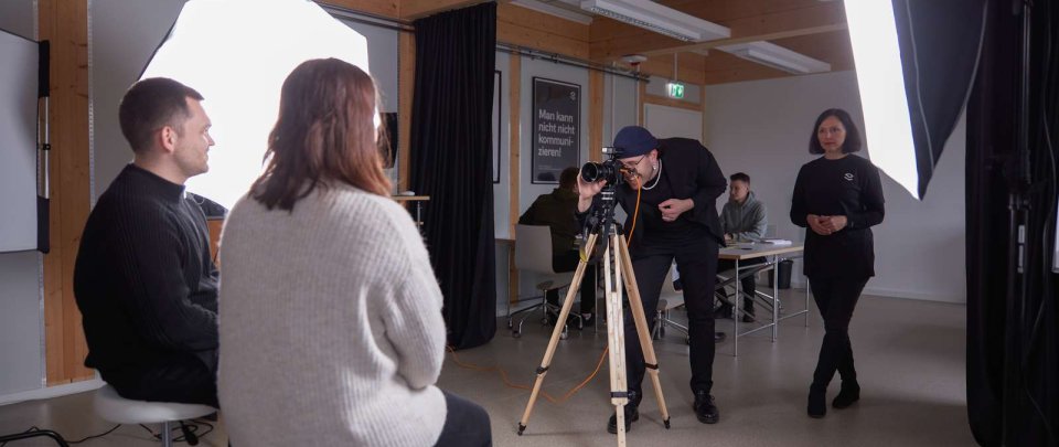 In einem Raum sitzen zwei Studierende auf Hockern und werden angestrahlt, ein weiterer Student macht mit einer Kamera auf einem Stativ Fotos von ihnen, während eine Dozentin in schwarzer Kleidung neben ihm steht.