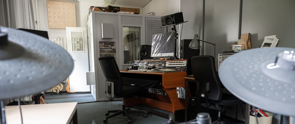 Ein Mischpult, davor unscharf Teile eines Schlagzeigs, dahinter eine Aufnahmekabine.