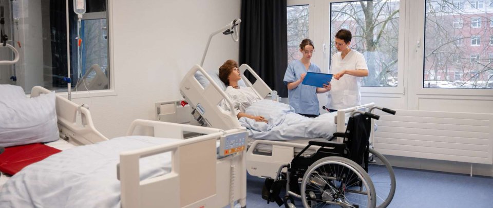 HealthLab der Hochschule Flensburg: Ein Raum, der eingerichtet ist wie ein Krankenhauszimmer. Eine Dozentin und eine Studentin stehen gemeinsam an einem Bett mit einer lebensgroßen Puppe.