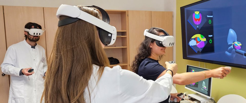 Zwei Frauen und ein Mann mit VR-Brillen stehen vor einem Monitor mit bunten Formen.