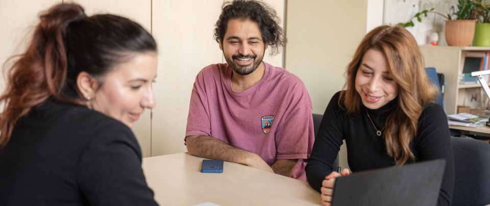 Three people sitting around a desk-like table. One of them seems to be showing the other two something on a laptop screen.