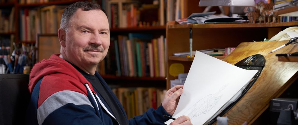 Andreas Deja, kurze Haare, lächelnd der Kameera zugewandt, in T-Shirt und Sweatjacke, an seinem Schreibtisch. Er hält Stift und Zeichenbogen.