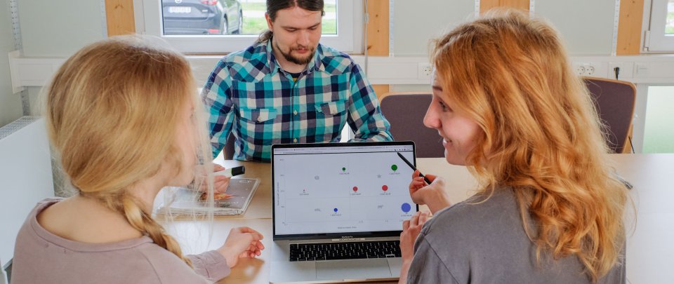 Zwei junge Frauen vor einem Laptop. Eine zeigt der anderen etwas. Im Hintergrund ein weiterer Mensch.