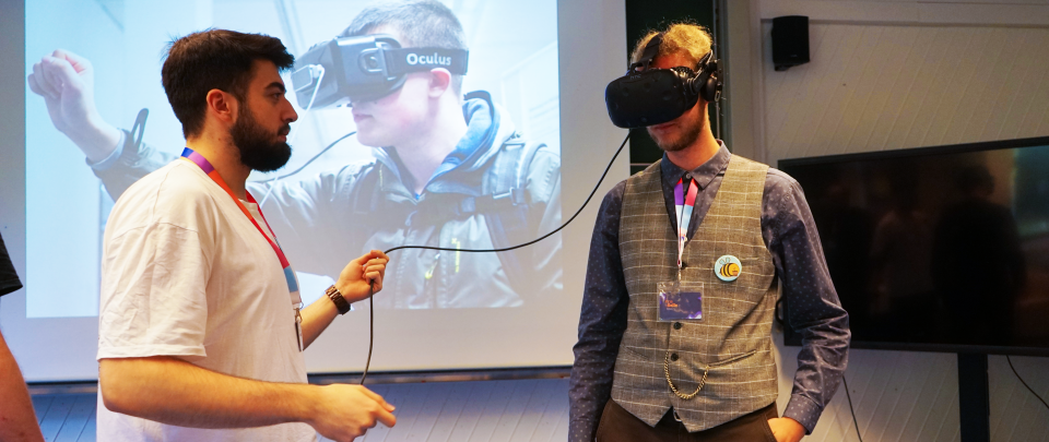 Zwei junge Männer, einer trägt eine VR-Brille, der andere hält ein Kabel, das mit der Brille verbunden ist.