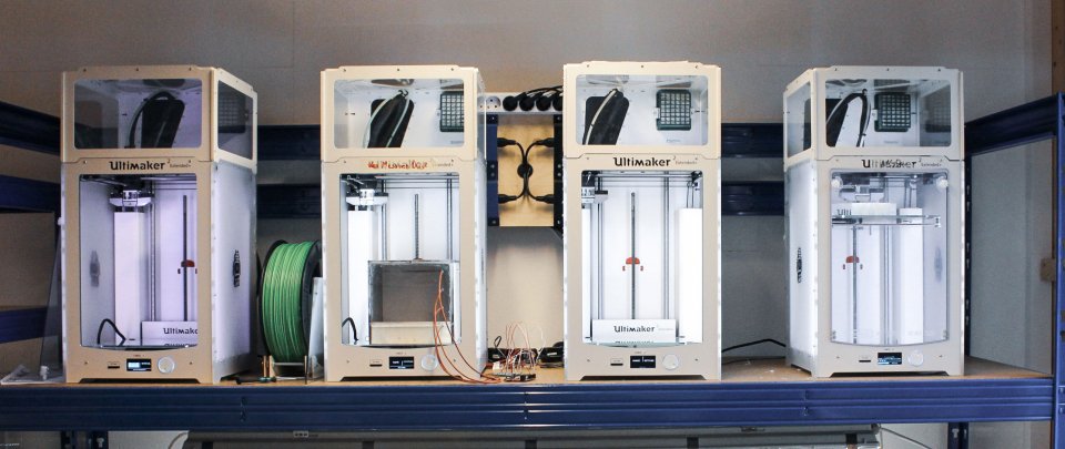 Auf einem Regal stehen nebeneinander vier 3D-Drucker.