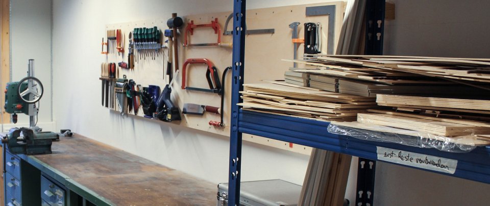 Blick in die analoge Werkstatt: Vorn rechts ein Regal mit Holzplatten, dahinter eine Werkbank und an der Wand darüber diverse Werkzeuge.