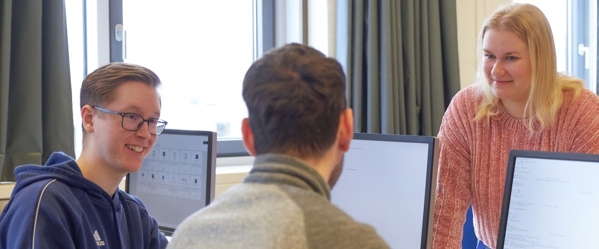 Studierende in einem Seminarraum. Zwei Studenten schauen auf Bildschirme, eine Studentin steht dahinter. Sie unterhalten sich.
