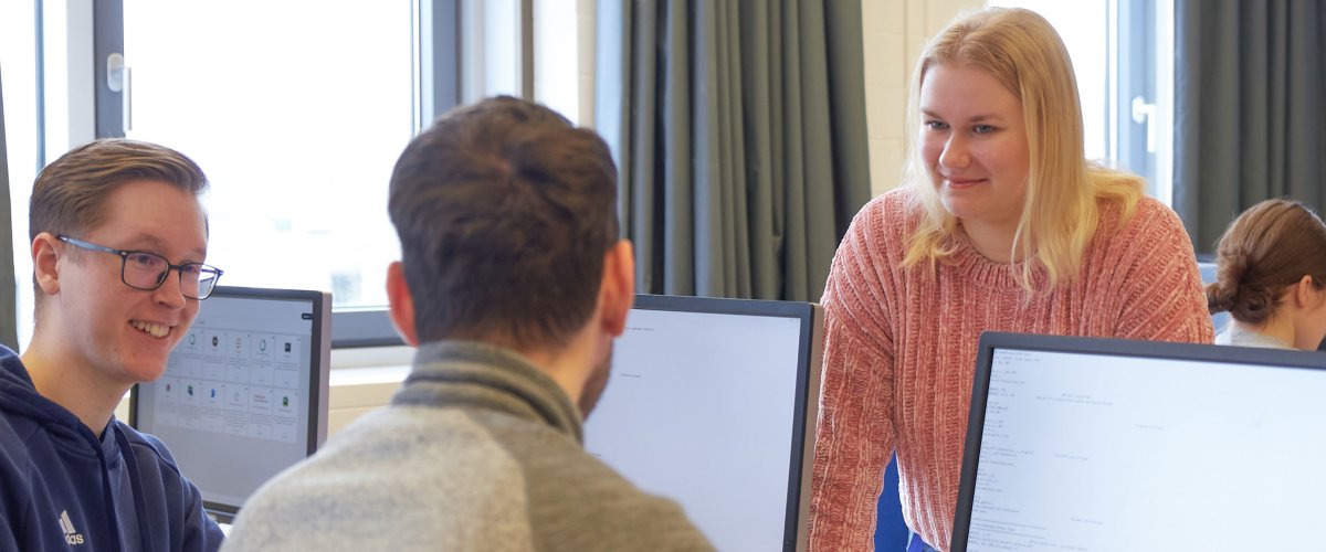 Studierende in einem Seminarraum. Zwei Studenten schauen auf Bildschirme, eine Studentin steht dahinter. Sie unterhalten sich.