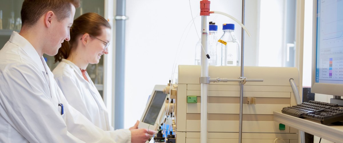 Zwei Studierende im Labor. Sie tragen Kittel und schauen auf ein Gerät und Bildschirm vor ihnen.