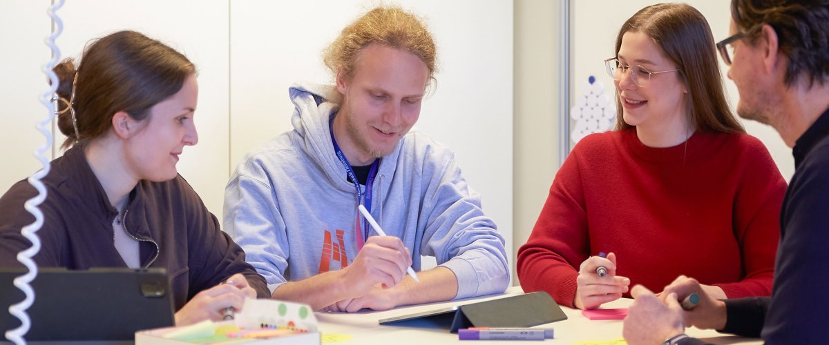 Studierende und ein Dozent stehen um einen Tisch. Sie halten Stifte, einer macht Notizen auf dem Tablet.