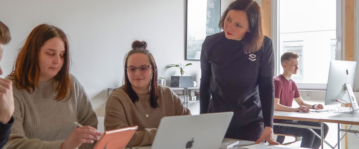 Studierende arbeiten in einem hellen, freundlichen Raum an Laptops. Eine DSozentin steht daneben und hört interessiert zu.