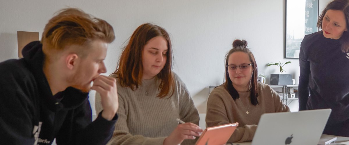 Studierende arbeiten in einem hellen, freundlichen Raum an Laptops. Eine DSozentin steht daneben und hört interessiert zu.