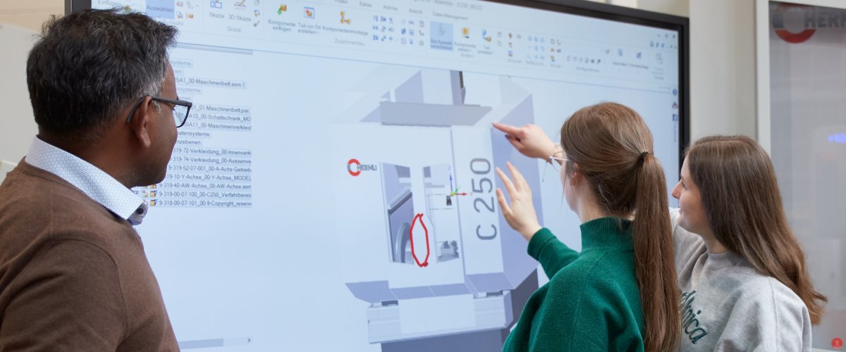 Zwei Studentinnen stehen an einem großen Monitor und zeigen auf die dort dargestellte Grafik mit verschiedenfarbigen eckigen Flächen und der Angabe C250. Ein Dozent steht links, ein Student links neben ihnen und blicken ebenfalls auf den Bildschirm.