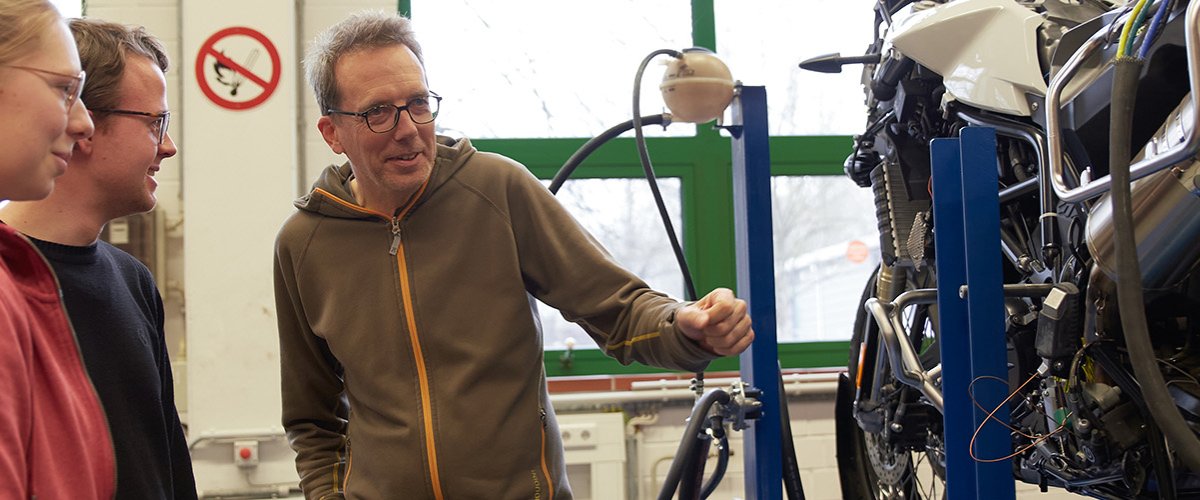 Zwei Studierende und ein Dozent stehen vor einem Motorrad.