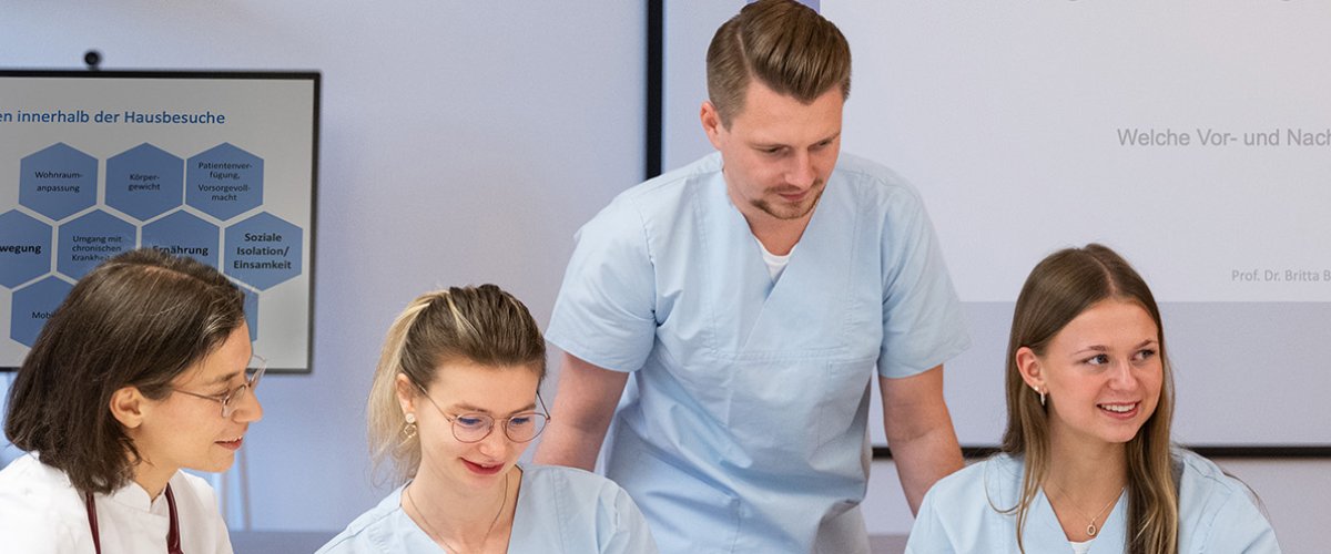 Eine Gruppe von drei Studierenden und zwei Dozentinnen sitzt an einem Tisch, im Hintergrund eine Präsentations-Projektion an der Wand und ein Monitor mit einem Diagramm. Alle tragen hellblaue Krankenhaus-Pfleger-Kittel.
