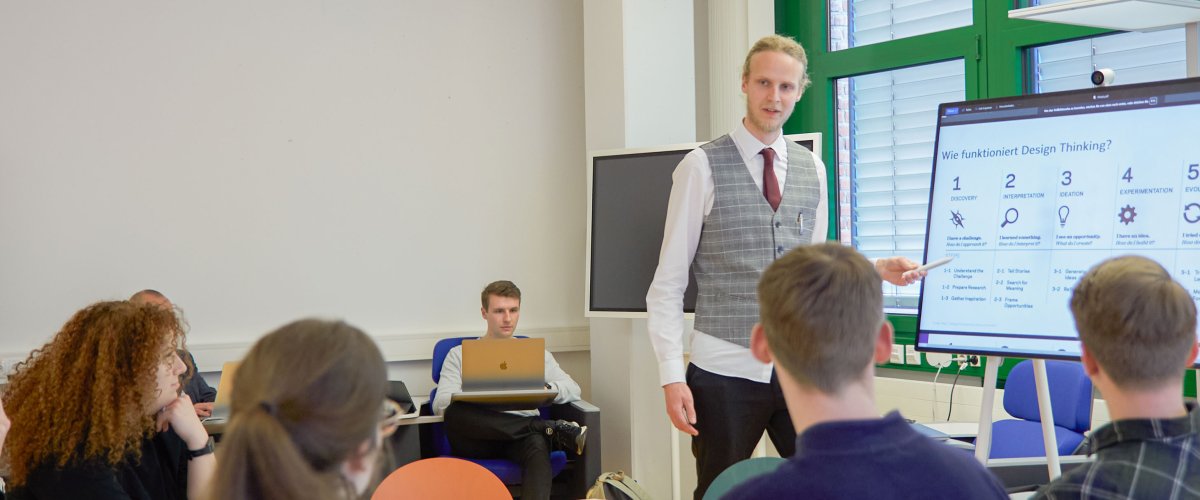 Seminarszene: Student steht neben Dozent an einem Bildschirm und erläutert etwas. Im Vordergrund andere Studierende um einen Tisch.