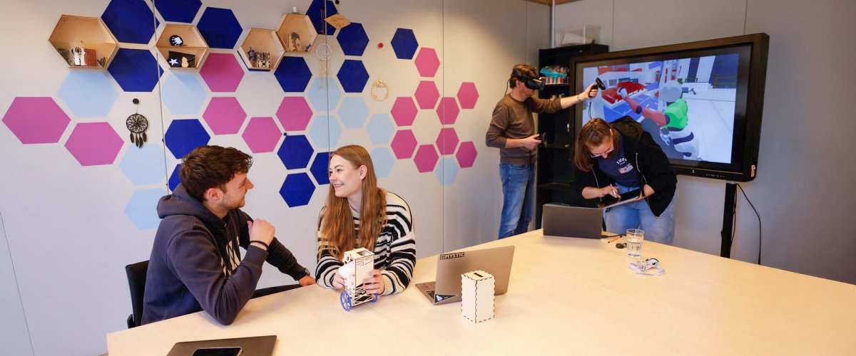 In einem Seminarraum mit bunten Kacheln an der Wand sitzen zwei Studierende an einem Tisch und unterhalten sich, auf dem Tisch vor ihnen steht ein kleiner Roboter. Im Hintergrund steht ein Dozent vor einem Monitor und trägt eine VR-Brille, neben ihm eine weitere Studentin, die etwas auf einem Tablet nachschaut.