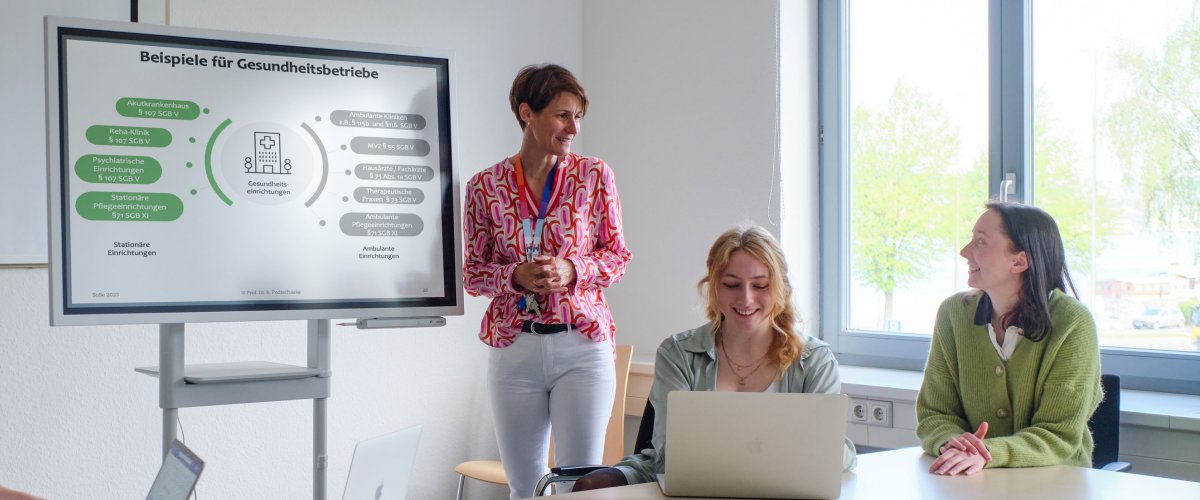 Seminarszene: Eine Dozentin steht vor einem Whiteboard, am Tisch sitzt eine Gruppe von vier Studierenden, die lächelnd auf ihre Laptops oder nach vorn zum Whiteboard schauen. Auf dem Whiteboard ist eine Grafik mit der Überschrift "Beispiele für Gesundheitsbetriebe" zu sehen.