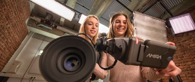 Zwei Studentinnen schauen auf das Display einer Kamera, die im Bildvordergrund zu sehen ist. Die Studentinnen sind im TV-Studio der Hochschule.