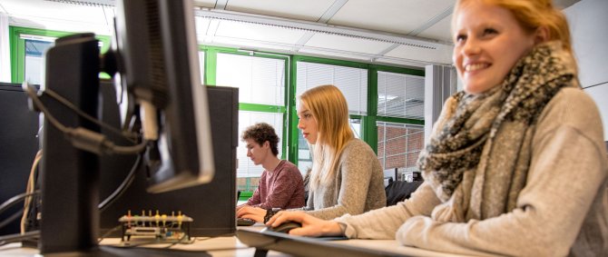 Drei Studierende arbeiten in einem Computerlabor je an einem Rechner. Vorn im Bild und am besten zu erkennen eine blonde lachende Frau.