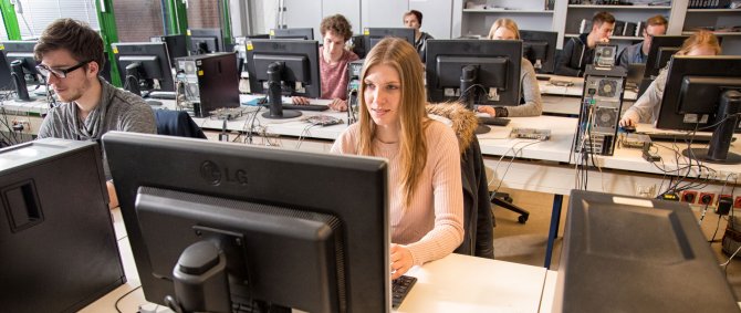 Blick auf ein Computerlabor, in dem mehrere Studierende an Rechnern arbeiten. In der ersten Reihe und am besten zu erkennen eine blonde Frau und ein dunkelhaariger Mann.